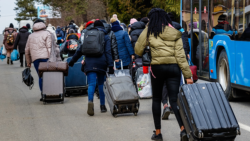 Flüchtlinge, die mit Koffern einen Weg entlang gehen, daneben ein Bus.