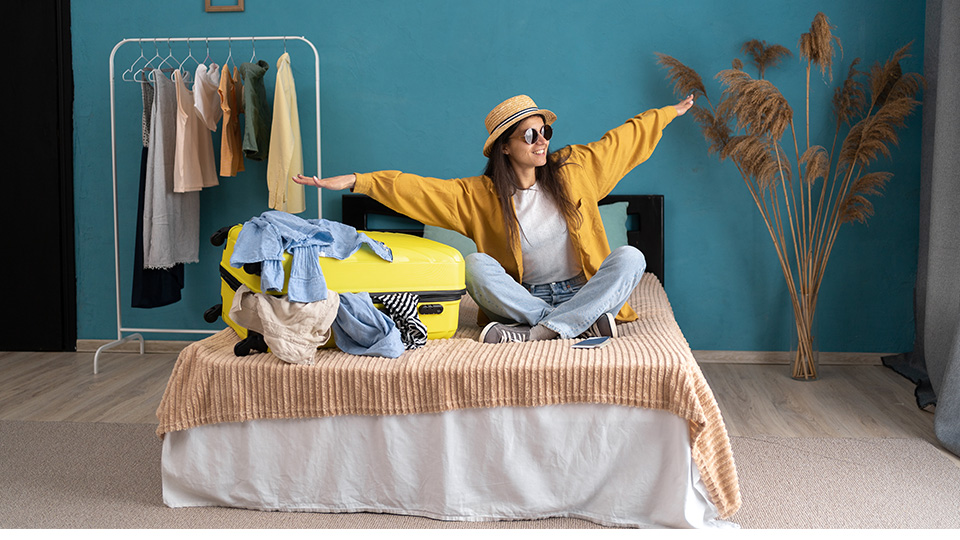 Ferien zuhause: Frau mit Sonnenbrille, die mit ihrem Koffer auf ihrem Bett sitzt und die Arme in die Luft streckt