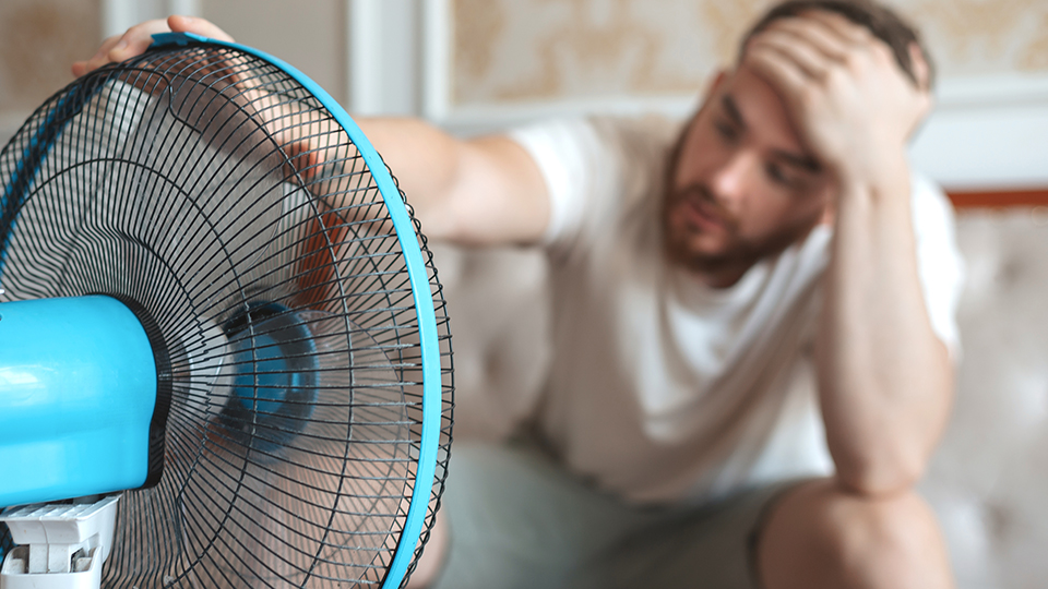 Steigende Temperaturen: Mann sitzt mit Hand auf der Stirn vor laufendem Ventilator