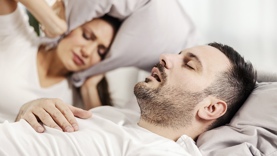 Schnarchen: Mann, der schnarcht und Frau, die sich die Ohren zuhält