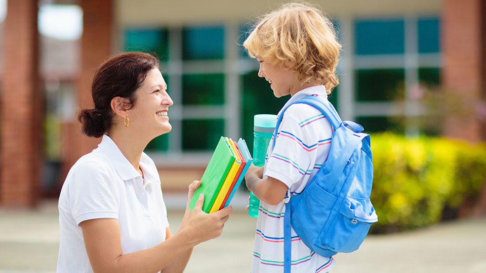 Mutter, die ihrem Kind am ersten Schultag Schulmaterialien in die Hand gibt und ihn anlächelt.