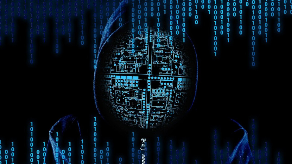 Cyber-Kriminalität: Vermummte Person vor Bildschirm mit Zahlencodes, dunkler Hintergrund