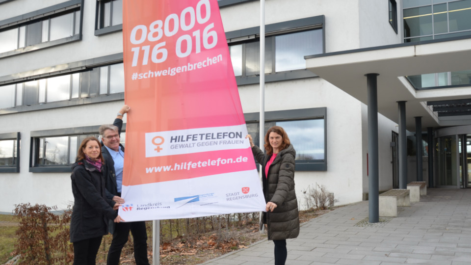 Landrätin Tanja Schweiger (rechts), Gleichstellungsbeauftragte Silvia Siegler und Markus Gschwendtner, Beauftragter für Chancengleichheit am Arbeitsmarkt des Jobcenters Landkreis Regensburg, hissen gemeinsam die Fahne des Hilfetelefons „Gewalt gegen Frauen“ im Landratsamt Regensburg.