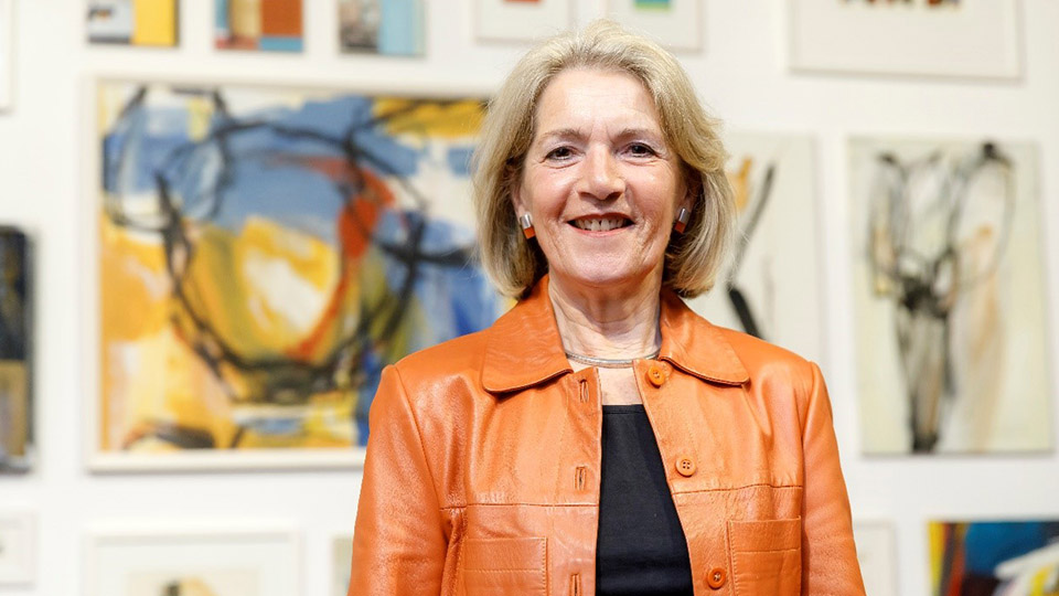 Künstlerin Maria Maier lächelt in die Kamera, hinter ihr eine Kunst-Ausstellung