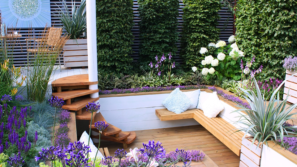 Garten mit Holzbank und im Hintergrund Holztisch und Stühlen, im Vordergrund lilafarbene Blumen.