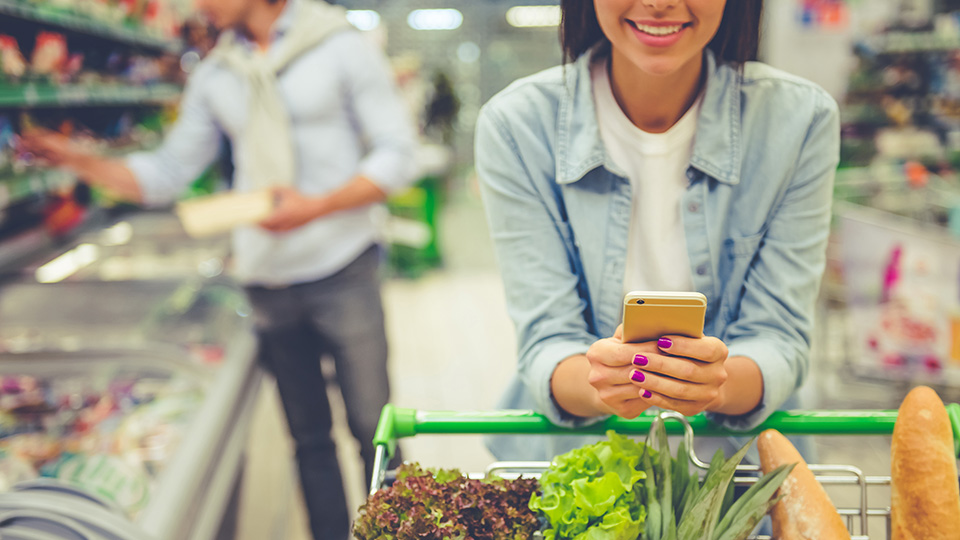 Frau schiebt Wagen mit Einkäufen, Freund holt im Hintergrund Lebensmittel aus dem Regal