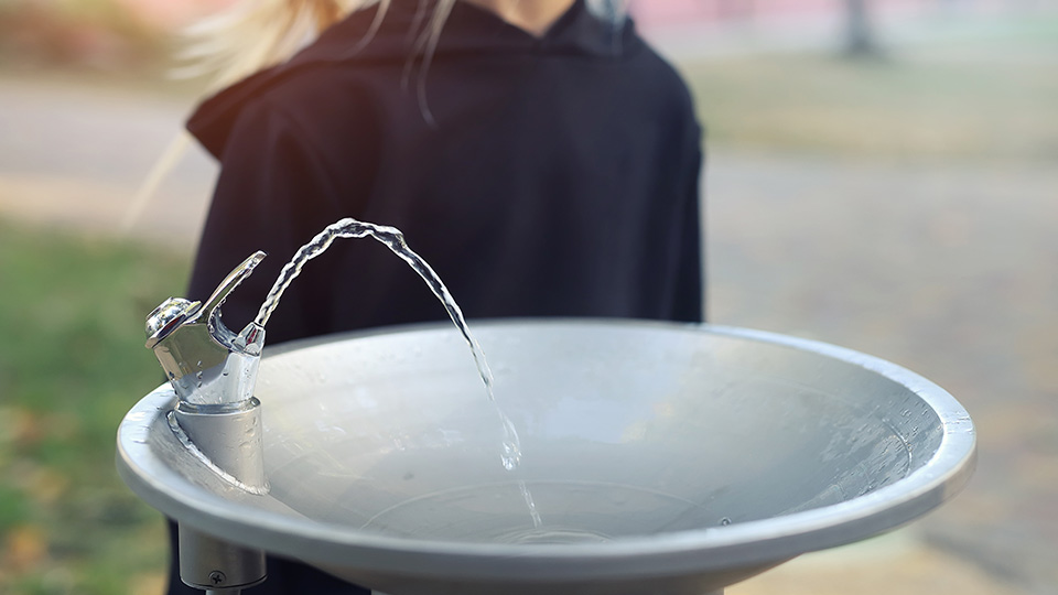 Trinkwasserbrunnen, der gerade läuft, dahinter ein blondes Mädchen.