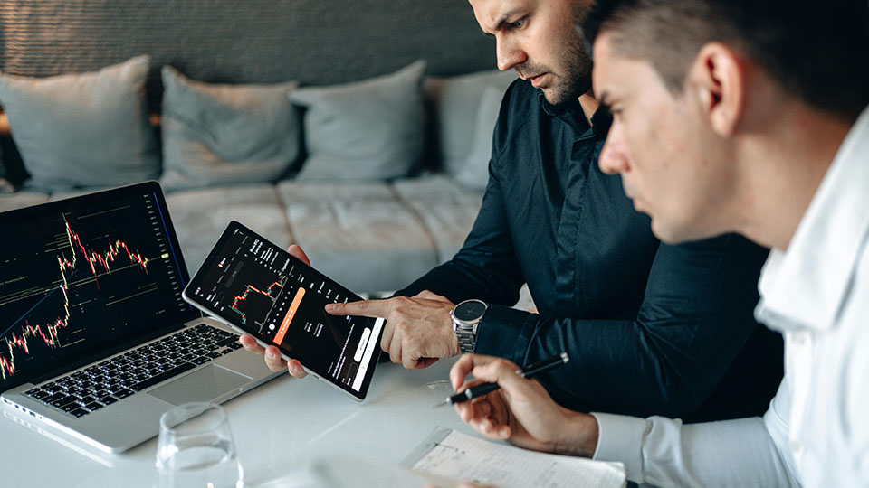 Zwei Männer schauen sich den Kurs von Kryptowährungen auf dem IPad und Laptop an