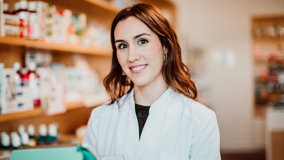 Frau mit weißem Kittel lächelt in die Kamera; im Hintergrund verschwommen Apotheken-Regale