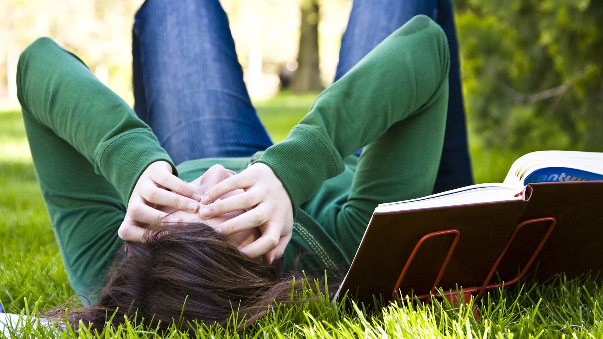 Gestresster Student liegt im Gras neben Buch und hält sich Hände vors Gesicht.
