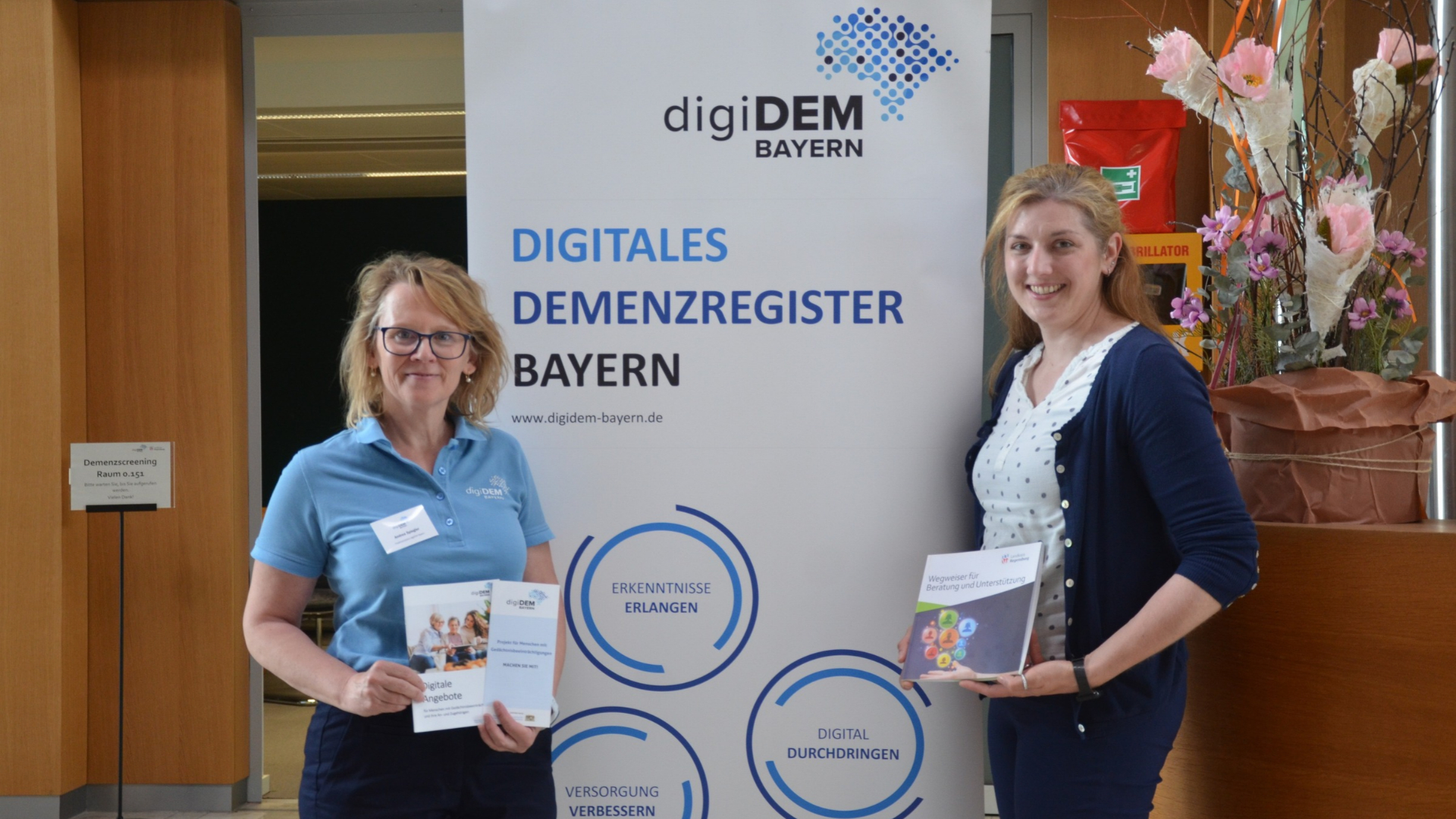 Zwei Damen stehen vor einem Roll-up Banner und präsentieren den Demez-Screeningtag des Landratsamts